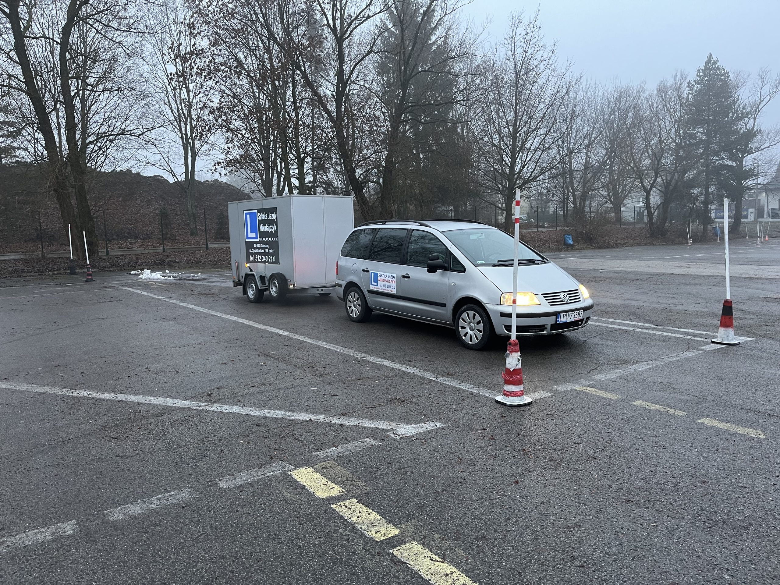 Srebrny samochód typu minivan z przyczepą stoi na placu manewrowym w mglisty dzień. Na aucie i przyczepie widoczne są oznaczenia nauki jazdy (litera "L"). Pojazd znajduje się między białymi słupkami wyznaczającymi tor jazdy.